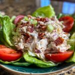 A serving of BLT Chicken Salad on a bed of lettuce with tomato wedges. Crispy bacon and green onions top the salad.