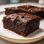 A close-up of flourless greek yogurt brownies on a white plate, topped with chocolate chips.
