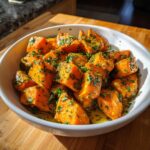 A bowl of Garlic Butter Sweet Potatoes, garnished with fresh herbs, sits on a wooden surface.