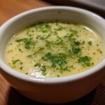A bowl of creamy Garlic Herb Potato Soup, garnished with fresh herbs, sits on a wooden surface.
