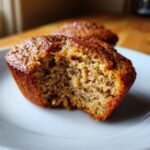 A partially eaten healthy banana muffin on a white plate, showcasing its moist texture.