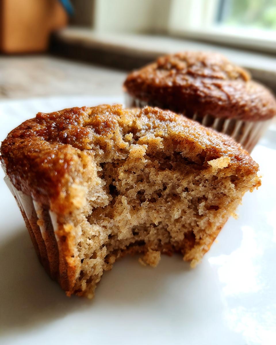 A close-up of a Healthy Banana Muffin with a bite taken out, showing the moist texture, with another muffin in the background.