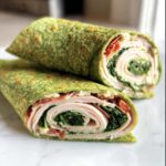 Two halves of a Keto Spinach Wrap filled with turkey, spinach, and bacon on a white surface.