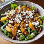 A bowl of Millionaire Peach Salad with fresh peaches, feta cheese, pecans, lettuce, and balsamic glaze.