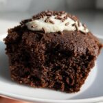 A close-up of a Protein Brownie topped with white cream and chocolate sprinkles on a white plate.