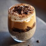 Close-up of a layered Viral Tiramisu Chia Pudding in a glass, topped with whipped cream and chocolate.
