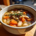 A bowl of hearty White Bean Soup with carrots, herbs, and a sprig of thyme on top.