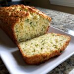 A loaf of Zucchini Cheddar Quick Bread with a slice cut, showcasing the zucchini and cheddar.