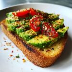 Close-up of Avocado Toast topped with sliced avocado, cherry tomatoes, and herbs on toasted bread.