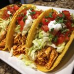 Three Easy Chicken Tacos on a white plate, filled with chicken, lettuce, tomato, and sour cream.