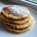 A stack of four golden Fluffy Pancakes from Scratch, dusted with powdered sugar on a white plate.