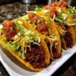 Close-up of several Ground Beef Tacos filled with meat, lettuce, cheese, and salsa on a white plate.