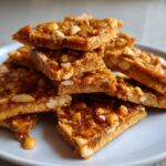 A stack of delicious Homemade Peanut Brittle pieces on a white plate, showcasing the peanuts and brittle texture.
