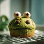 A green Monster Muffin for Kids decorated with candy eyes and chocolate chips on a wire rack.