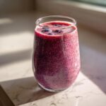 A glass filled with a purple breakfast smoothie, topped with blueberries, sits on a marble surface.