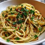 Close-up of Spaghetti Aglio e Olio in a white bowl, garnished with fresh parsley and red pepper flakes.
