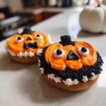 Two spooky Halloween bagel ideas, decorated as pumpkins with orange frosting, black sprinkles, and candy eyes.
