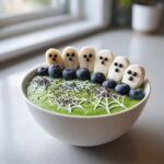 A Spooky Halloween Smoothie Bowl with green smoothie, banana ghosts, blueberries, and a spiderweb design.