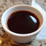 A close-up of The Classic Au Jus, a rich, dark brown liquid in a white ramekin, ready to enhance any dish.