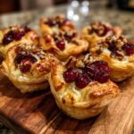 Close-up of golden-brown Cranberry Brie Cups topped with cranberries and walnuts on a wooden board.