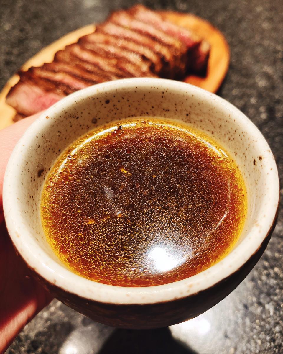 Close-up of a bowl of rich, flavorful Au Jus, with sliced steak in the background.