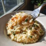 A fork lifts a cheesy bite of creamy Parmesan Risotto, topped with grated cheese and herbs.