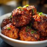 A close-up of a bowl filled with glistening, glazed Party Meatballs, garnished with fresh herbs.