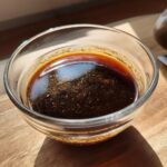 A clear glass bowl filled with rich, dark homemade au jus, with small particles of herbs and spices visible.