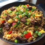 A close-up of a pan filled with savory Prime Rib Fried Rice, featuring tender beef, scrambled eggs, red peppers, and green onions.