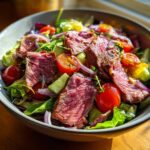 A vibrant bowl of Prime Rib Salad (Light) featuring sliced medium-rare prime rib, cherry tomatoes, cucumber, and red onion on a bed of mixed greens.