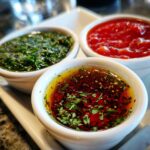 Three small white bowls filled with vibrant dips: a green herb dip, a red sauce, and an oil-based dip with spices and herbs, part of a Sparkling Dip Trio.