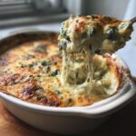 A scoop of cheesy, gooey baked Spinach Artichoke Dip being lifted from a white dish.