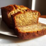 A freshly baked loaf of Brown Butter Banana Bread, with one thick slice cut and resting against the main loaf on a white plate.