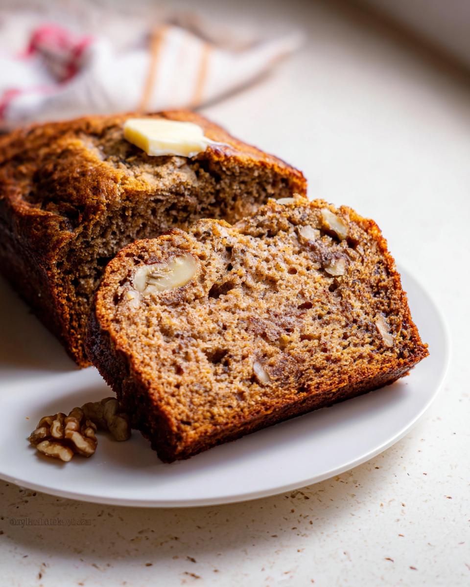 A thick slice of the Best Banana Bread Ever showing walnuts, served with the loaf and a pat of melting butter.