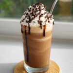 A tall glass of blended Mocha Frappuccino topped with whipped cream, chocolate syrup drizzle, and mini chocolate chips.