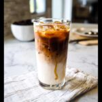 A tall glass showing the distinct layers of a Café-Style Iced Caramel Macchiato with ice, milk, and caramel drizzle.