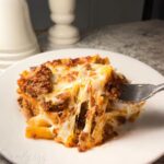 A fork pulls a cheesy, saucy bite from a serving of Baked Ziti for a Crowd, showing long cheese pulls.