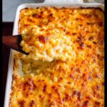 A wooden spoon lifting a generous scoop of creamy, cheesy Baked Mac and Cheese from a white baking dish.
