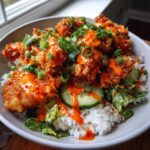 A close-up of Crispy Bang Bang Chicken Bowls featuring crispy fried chicken drizzled with orange sauce over rice, lettuce, and cucumber.