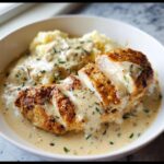 A sliced, pan-seared chicken breast covered in creamy ranch sauce, served alongside mashed potatoes.