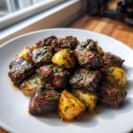 Close-up of Irresistible Garlic Butter Steak Bites & Potatoes coated in herbs and sauce on a white plate.