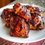 A plate piled high with juicy, caramelized BBQ Chicken Thighs featuring a dark, sticky glaze and char marks.