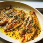 Close-up of three pan-seared Lemon Garlic Tilapia fillets swimming in a rich lemon-garlic butter sauce and topped with fresh parsley.