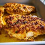 Close-up of golden, crispy breaded Melt In Your Mouth Chicken breasts sitting in buttery sauce.