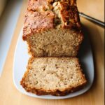Cross-section of a loaf of moist Vegan Banana Bread (Healthy) topped with walnuts, sliced on a white plate.