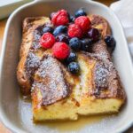 Close-up of a serving of Overnight French Toast Bake, topped with fresh raspberries, blueberries, and powdered sugar.