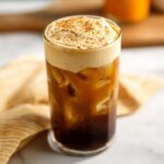 A tall glass filled with ice and dark cold brew coffee, topped with thick pumpkin cream foam dusted with spice, resembling Pumpkin Cream Cold Brew (Copycat).