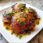 Close-up of three glistening Soy-Ginger Sticky Thighs coated in a dark glaze and topped with fresh green onions.