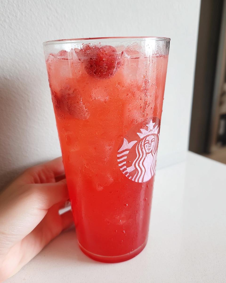 A cold, condensation-covered glass holding a vibrant red Strawberry Açaí Refresher Copycat with ice and strawberries.