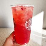A hand holds a clear plastic cup filled with a bright red, icy Strawberry Açaí Refresher Copycat drink.